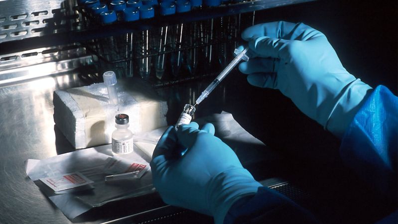 The image depicts a laboratory scene where a healthcare professional in blue gloves prepares a syringe with a vaccine from a vial. The setting is illuminated with focused lighting, highlighting the meticulous process of vaccine preparation.