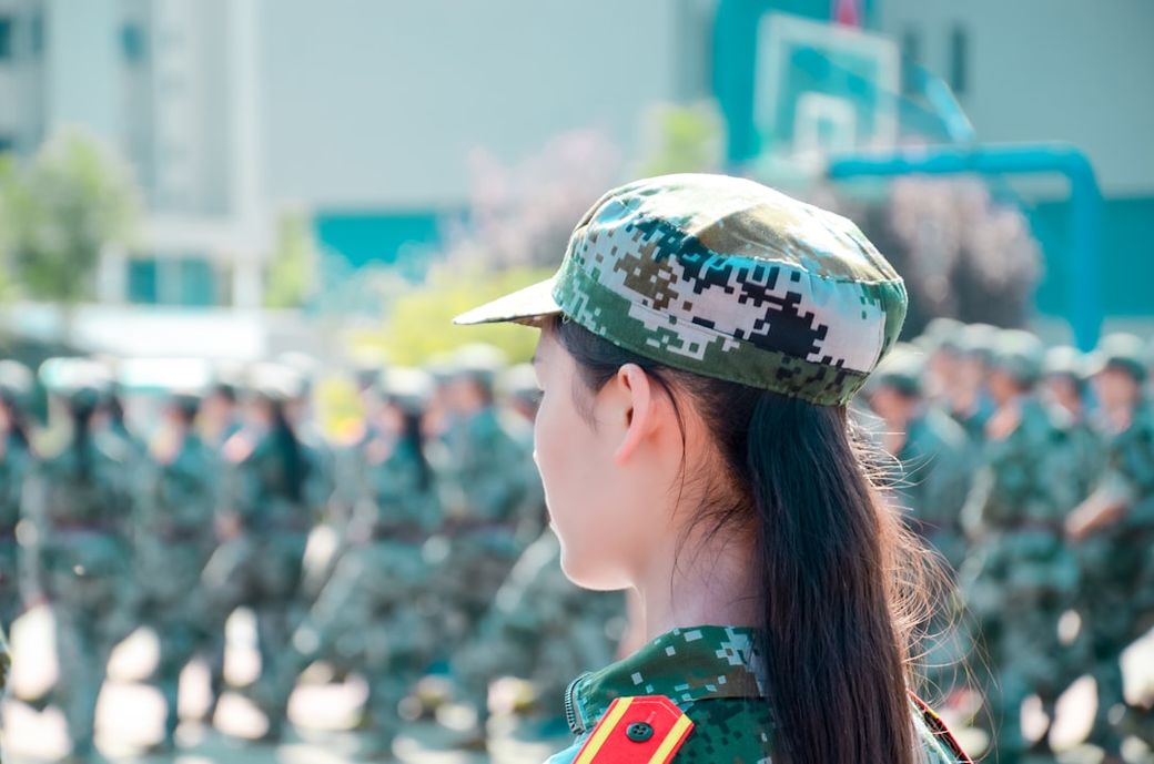 The image depicts a female soldier in uniform, wearing a camouflage hat, observing a group of soldiers participating in a drill or parade. The setting suggests a military training environment with a focus on discipline and organization. — Credit: Photo by Jerry Zhang on Unsplash