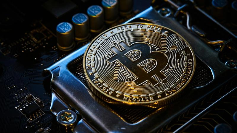 The image features a close-up of a Bitcoin, a cryptocurrency, resting on a metallic surface that resembles a computer processor. The intricate design of the Bitcoin showcases its digital nature, set against electronic components.