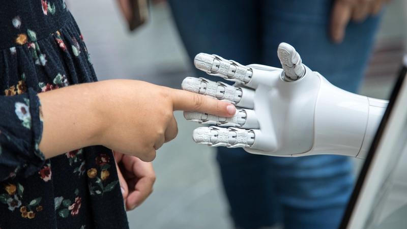 A child interacts with a robotic hand in a technological setting. The child reaches out to touch the robotic fingers, highlighting the connection between humans and advanced robotics.