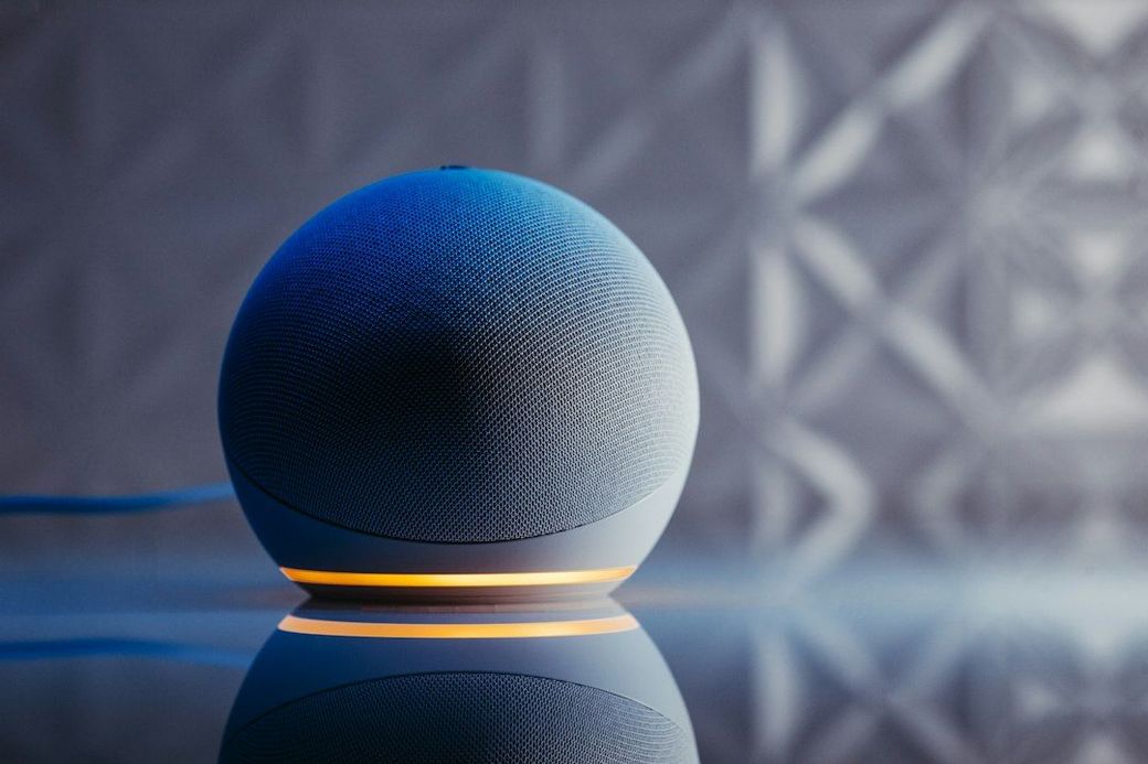 A close-up image of a smart speaker device featuring a spherical design and a glowing base. The background is softly illuminated with a geometric pattern, creating a modern and sleek ambiance. — Credit: Photo by Jonathan Borba on Unsplash