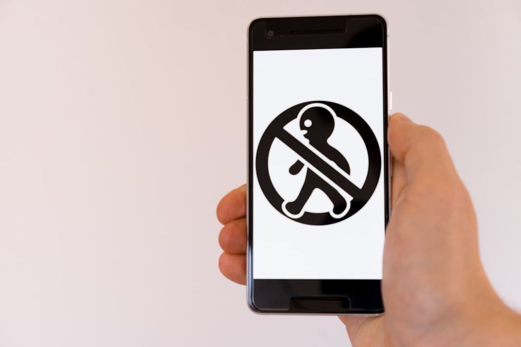 A person holds a smartphone displaying a no-entry sign featuring an abstract figure. The backdrop is plain, emphasizing the symbol on the screen. — Credit: Photo by Markus Winkler on Unsplash