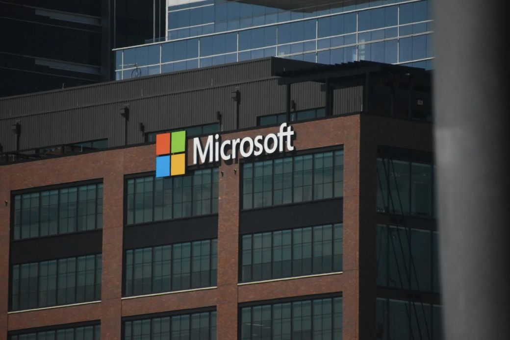The image features the exterior of a commercial building prominently displaying the Microsoft logo. The structure is modern with large windows and a distinctive facade. — Credit: Photo by Simon Ray / Unsplash