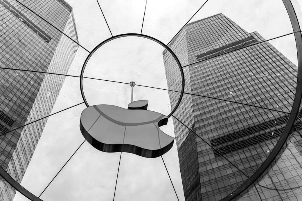 The image shows the iconic Apple logo suspended in an urban environment, framed by two tall glass skyscrapers. The black and white aesthetic enhances the modern architecture surrounding the logo. — Credit: Photo by Declan Sun on Unsplash