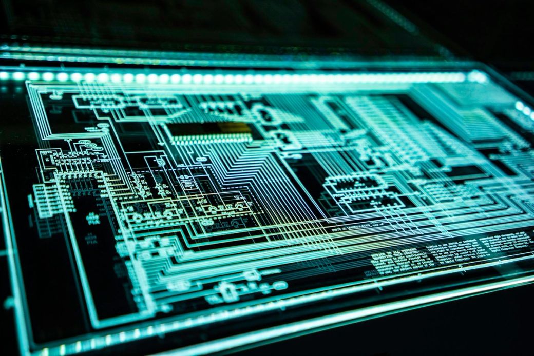 The image depicts a close-up view of a circuit board with intricate electronic pathways and components illuminated in a glowing blue light. This visually striking representation highlights the complexity and design of modern electronics. — Credit: Photo by Adi Goldstein / Unsplash