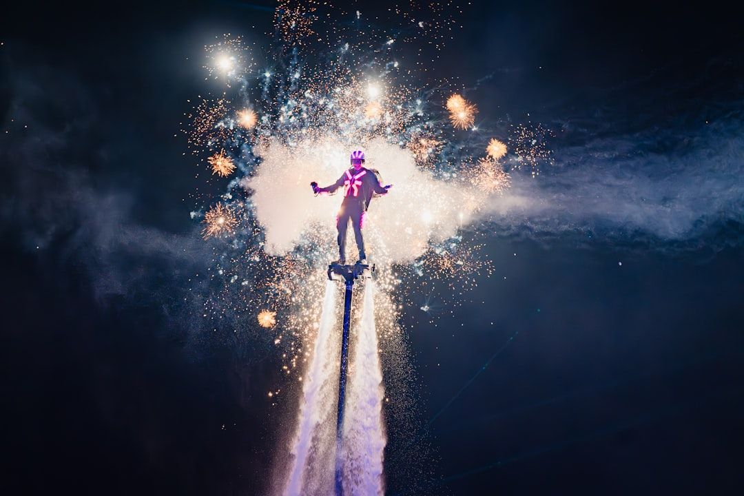 A performer is elevated above the ground on a hydroflighting device, surrounded by bursts of fireworks and vibrant lights. The scene captures a moment of excitement and entertainment at a night event.
