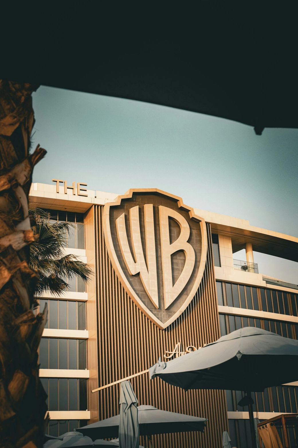 The image features the distinctive logo of Warner Bros. prominently displayed on a modern building facade. The architectural design is sleek and contemporary, surrounded by palm trees, suggesting a warm climate. — Credit: Photo by Dmitry Kropachev / Unsplash