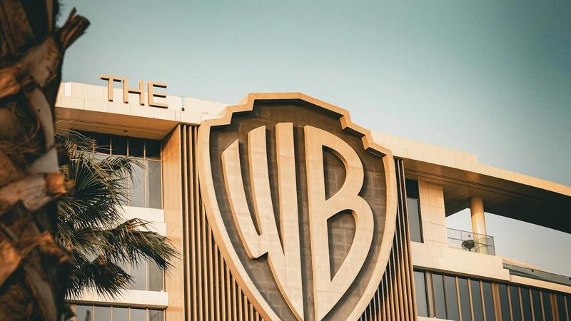 The image features the distinctive logo of Warner Bros. prominently displayed on a modern building facade. The architectural design is sleek and contemporary, surrounded by palm trees, suggesting a warm climate.