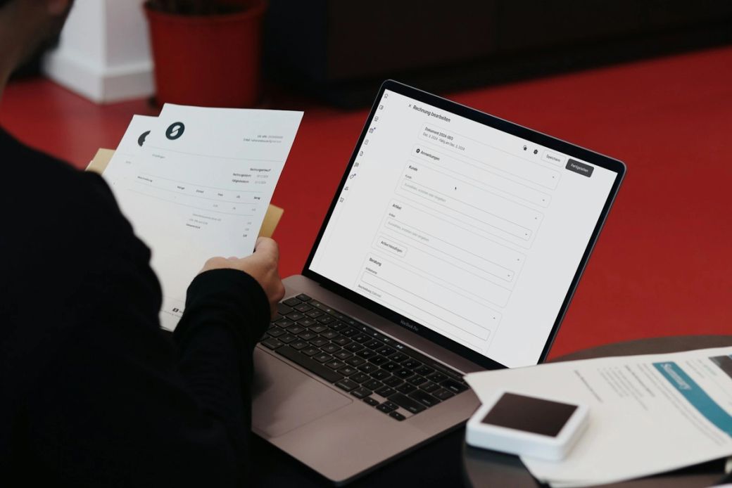A person is seated at a table reviewing documents while interacting with a laptop that displays an online form. The background features a red floor, creating a modern and casual setting. — Credit: Photo by SumUp / Unsplash