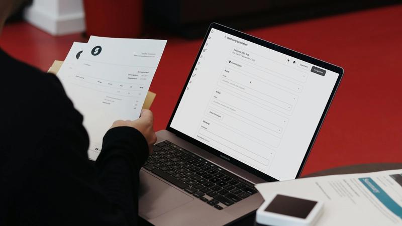 A person is seated at a table reviewing documents while interacting with a laptop that displays an online form. The background features a red floor, creating a modern and casual setting.
