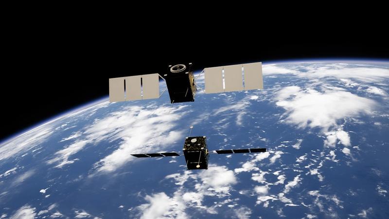 The image depicts two satellites in low Earth orbit, set against the backdrop of the planet's surface and cloud cover. The satellite structures are visible, showcasing their solar panels and technology.