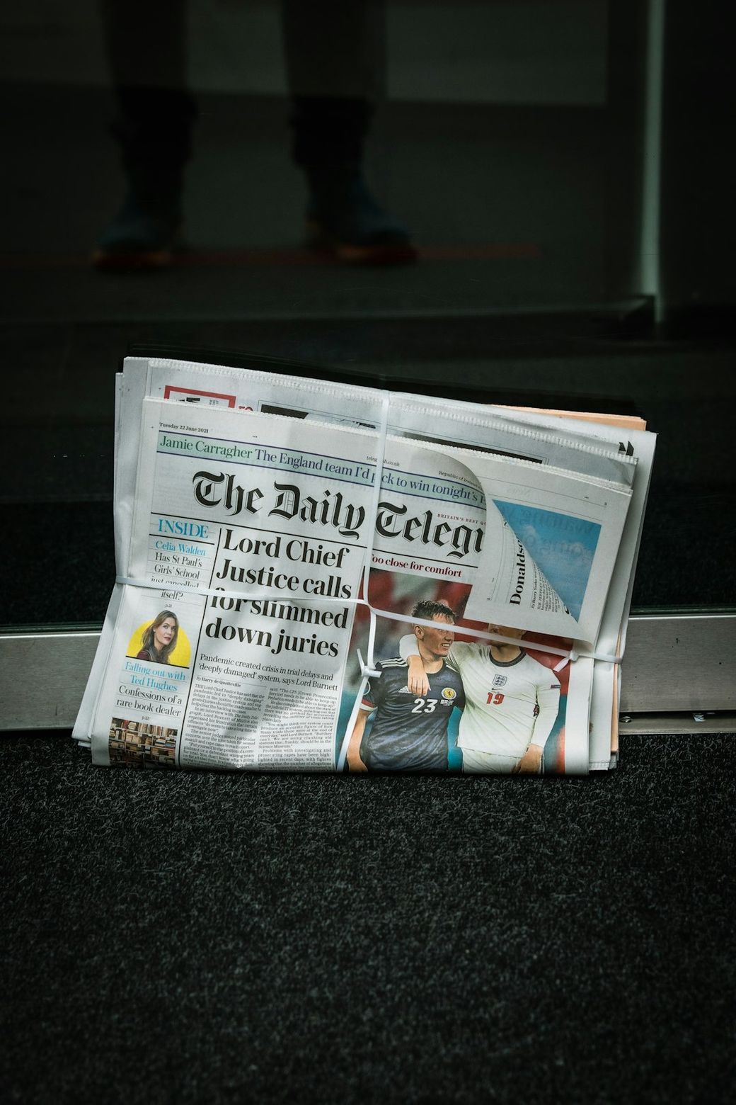 The image shows a folded newspaper titled 'The Daily Telegraph' resting on a floor near a doorway. The front page features a headline about a judicial issue and an article related to sports, indicating a blend of current news topics. — Credit: Photo by Kevin Grieve on Unsplash