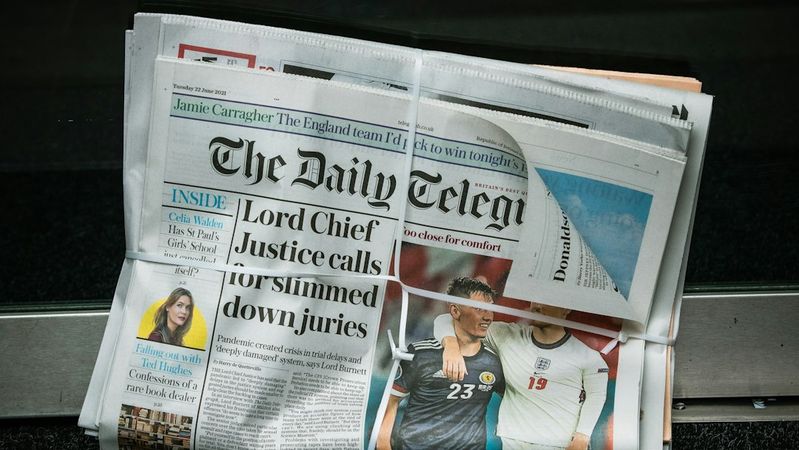 The image shows a folded newspaper titled 'The Daily Telegraph' resting on a floor near a doorway. The front page features a headline about a judicial issue and an article related to sports, indicating a blend of current news topics.