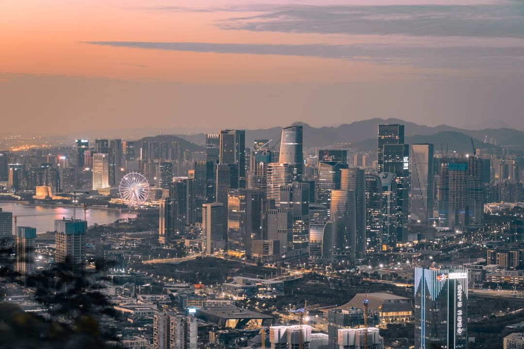 The image showcases a panoramic view of a modern city skyline at sunset, highlighting tall skyscrapers and a waterfront with a ferris wheel. The skyline is illuminated by city lights as dusk approaches.