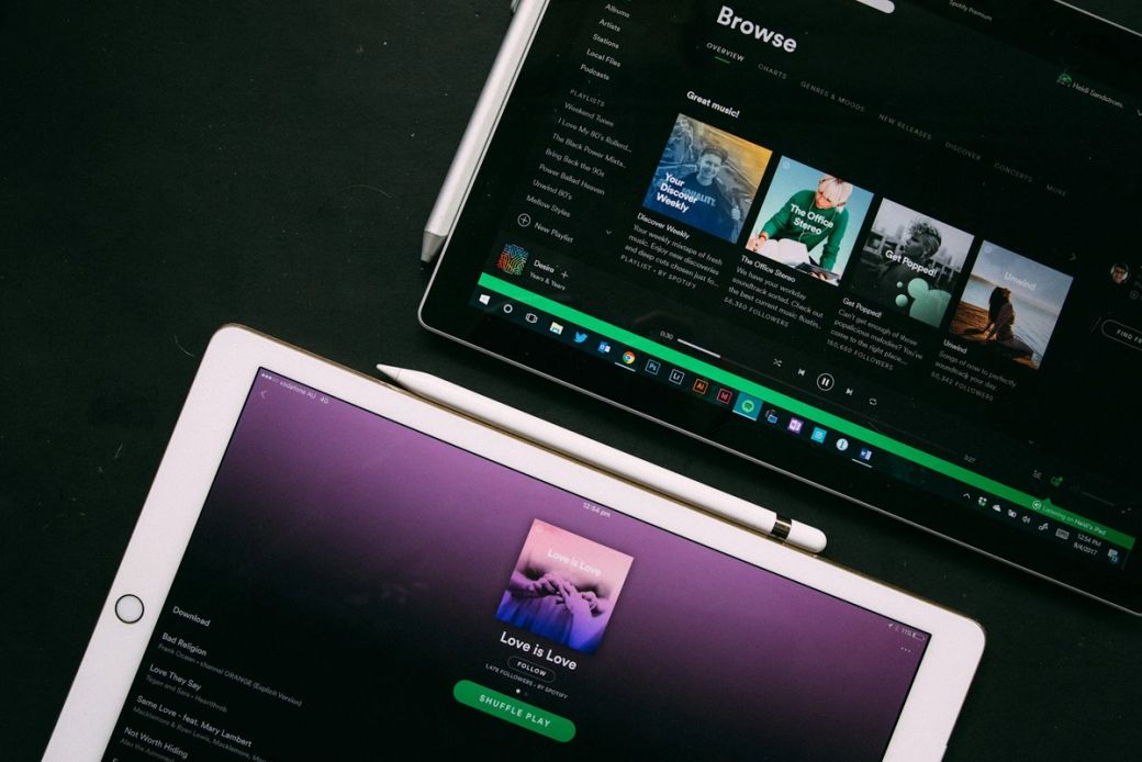 An overhead view of two tablets displaying music streaming applications. One tablet shows a playlist and album artworks on a dark background, while the other features an album titled 'Love is Love' against a purple backdrop. — Credit: Photo by Heidi Fin / Unsplash