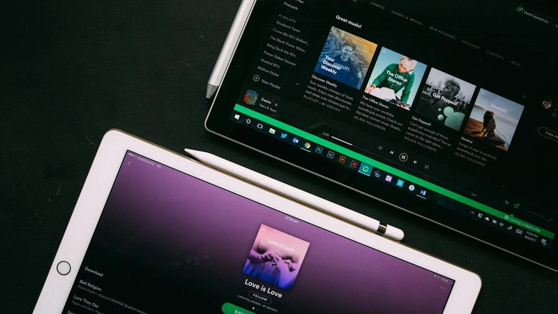 An overhead view of two tablets displaying music streaming applications. One tablet shows a playlist and album artworks on a dark background, while the other features an album titled 'Love is Love' against a purple backdrop.