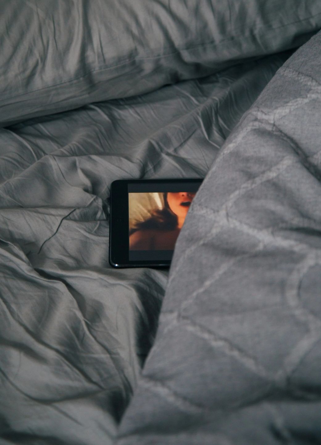 The image depicts a close-up view of a tablet resting on a bed, partially covered by a grey blanket. The screen shows an indistinct video content, suggesting a relaxed or private setting. — Credit: Photo by charlesdeluvio / Unsplash