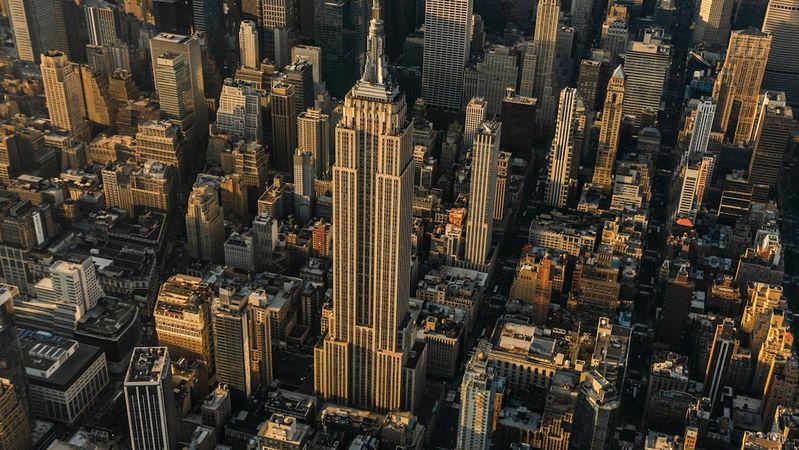 Aerial view of New York City with a focus on the iconic Empire State Building. The image captures the dense urban landscape, showcasing skyscrapers and the outline of the Hudson River in the background during sunset.
