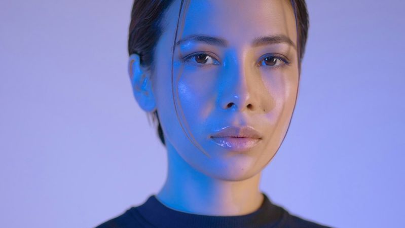 A close-up portrait of a young woman with slicked-back hair, set against a softly lit background illuminated in blue tones. The woman's expression is calm and introspective.