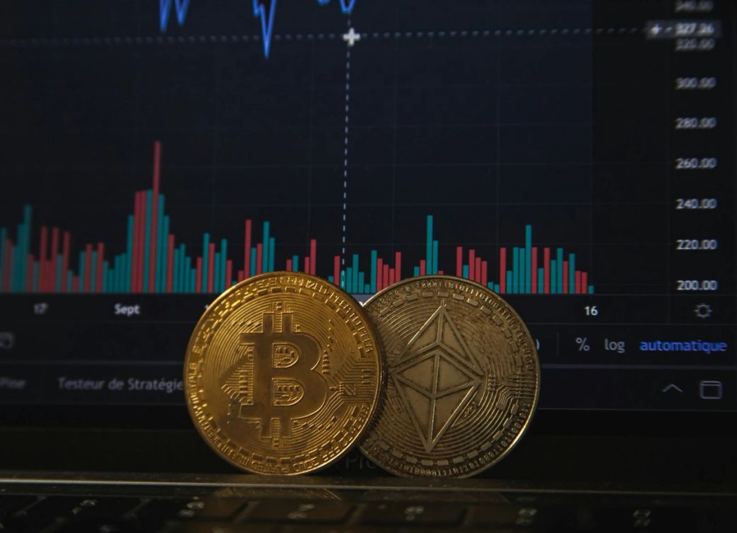 The image features two cryptocurrency coins, Bitcoin and Ethereum, placed in front of a digital financial chart displaying price fluctuations. The background shows a trading interface with colorful bar graphs, suggesting a focus on cryptocurrency markets. — Credit: Photo by Pierre Borthiry - Peiobty / Unsplash