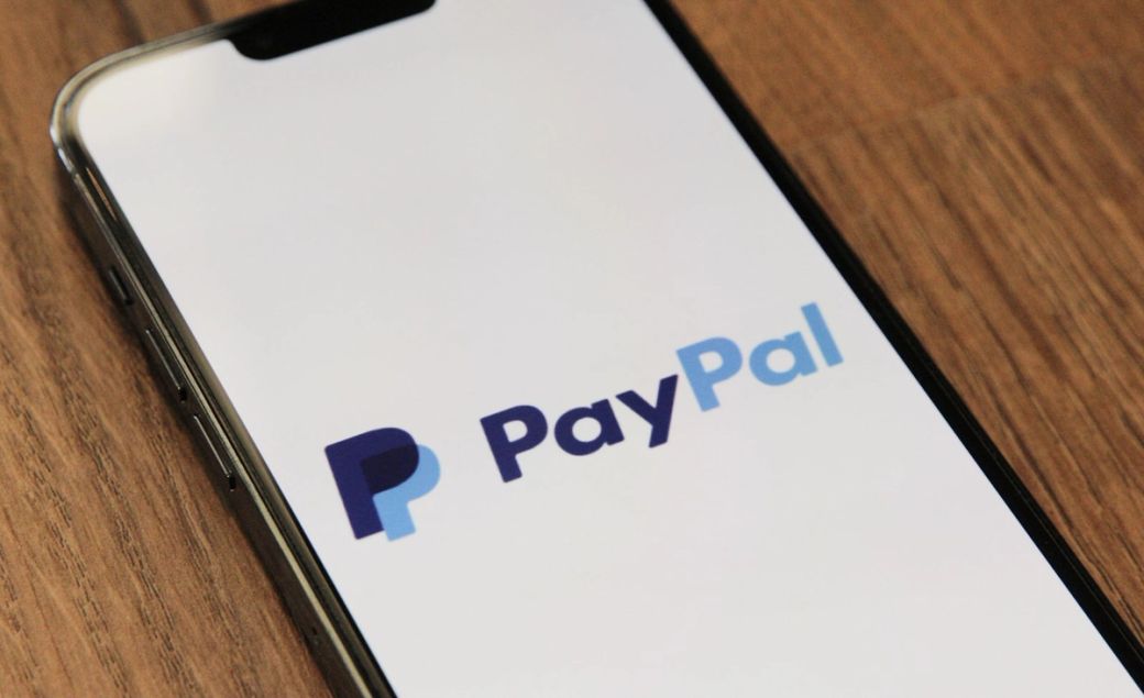 The image features a close-up view of a smartphone displaying the PayPal logo against a neutral background. The logo combines various shades of blue and is prominently placed on the device's screen. — Credit: Photo by Marques Thomas / Unsplash