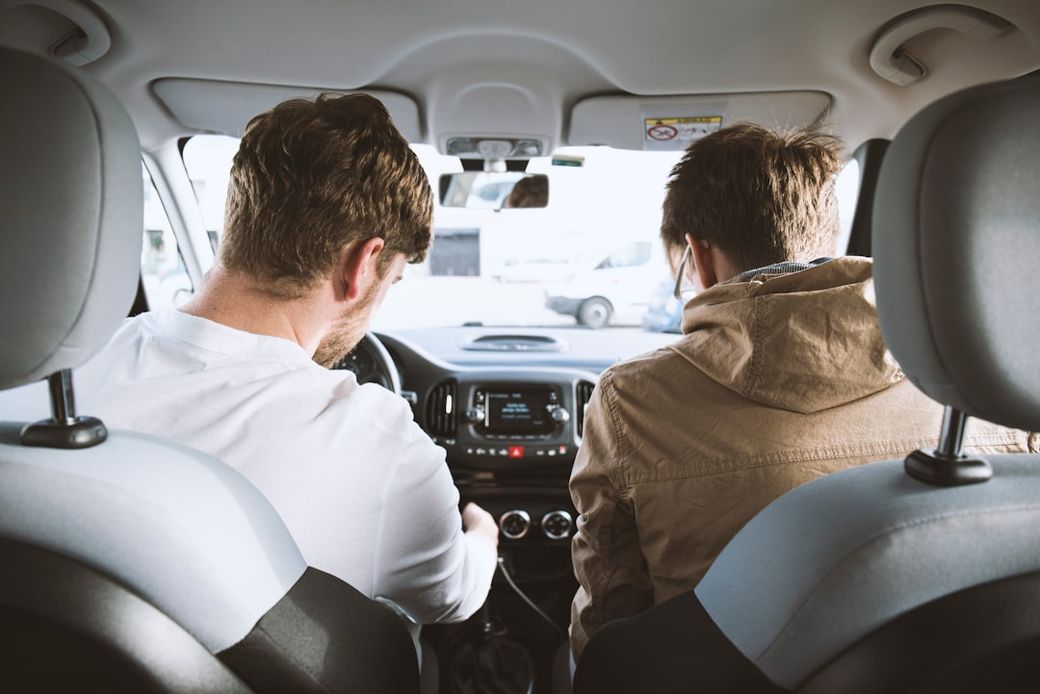 Two men are seated inside a car, one appears to be driving while the other is looking at the dashboard controls. The setting suggests a casual outing or a driving lesson. — Credit: Photo by David Emrich on Unsplash