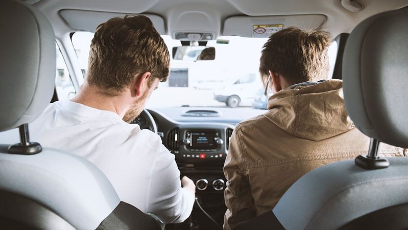 Two men are seated inside a car, one appears to be driving while the other is looking at the dashboard controls. The setting suggests a casual outing or a driving lesson.