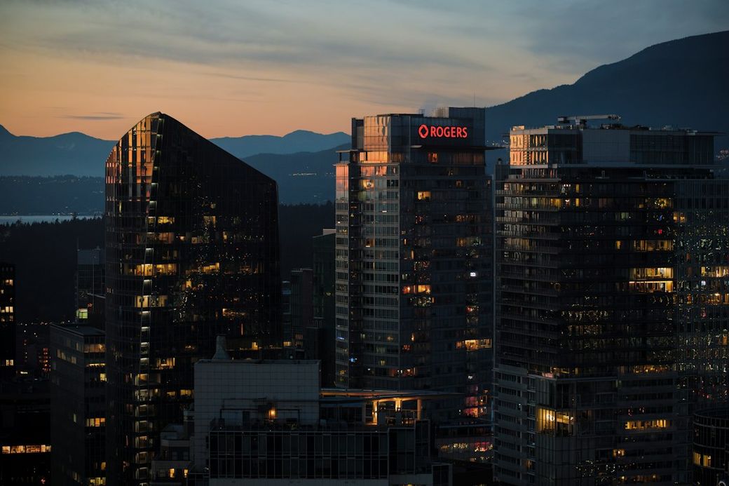 The image showcases a city skyline at dusk, featuring several high-rise buildings with illuminated windows. A prominent building displays the 'Rogers' logo against the backdrop of a colorful sunset sky and distant mountains. — Credit: Photo by Albert Stoynov on Unsplash