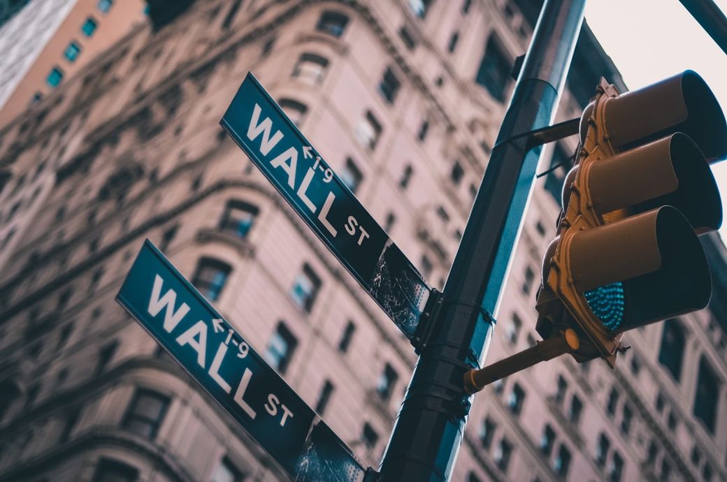 The image depicts a street sign for Wall Street against a backdrop of towering buildings in a financial district. The vantage point is from below, looking up at the signs, capturing the essence of the bustling financial hub. — Credit: Photo by Lo Lo on Unsplash