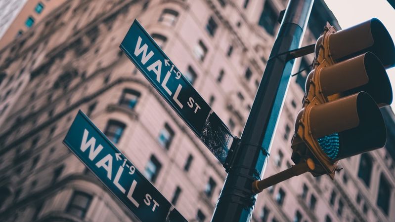 The image depicts a street sign for Wall Street against a backdrop of towering buildings in a financial district. The vantage point is from below, looking up at the signs, capturing the essence of the bustling financial hub.