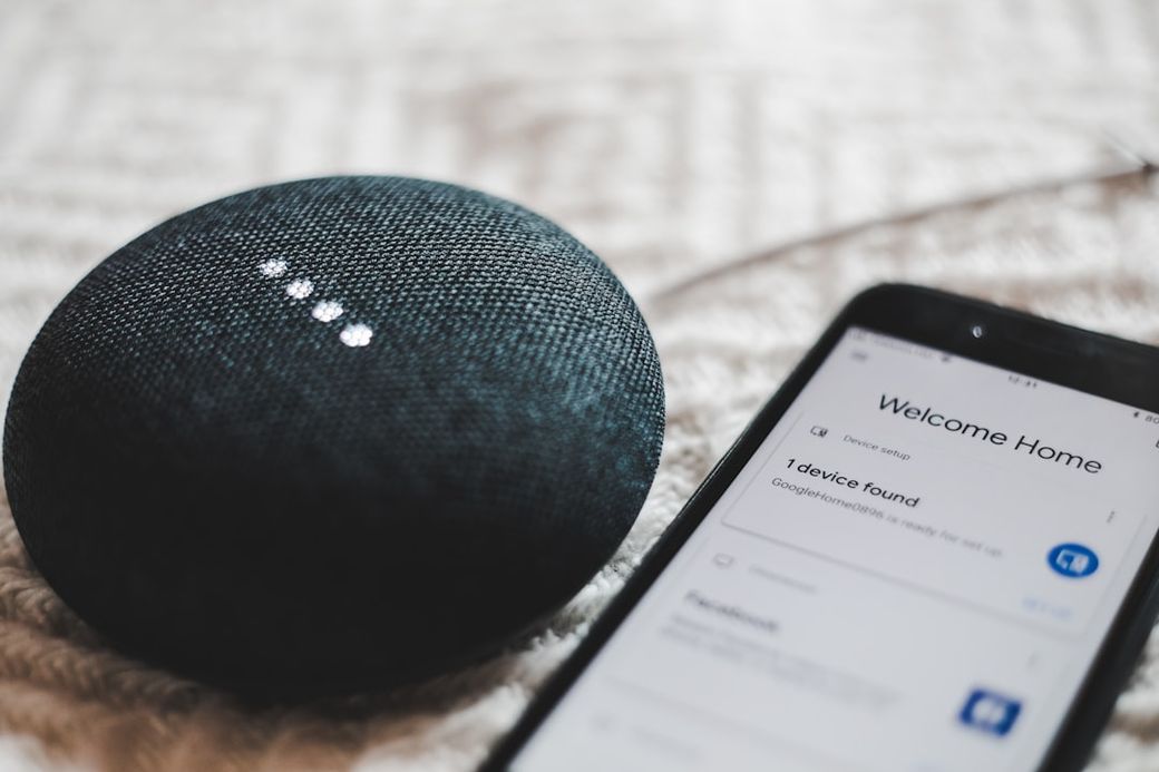 The image shows a black smart speaker sitting next to a smartphone on a textured surface. The smartphone screen displays a welcome message indicating that a device has been found. — Credit: Photo by BENCE BOROS on Unsplash