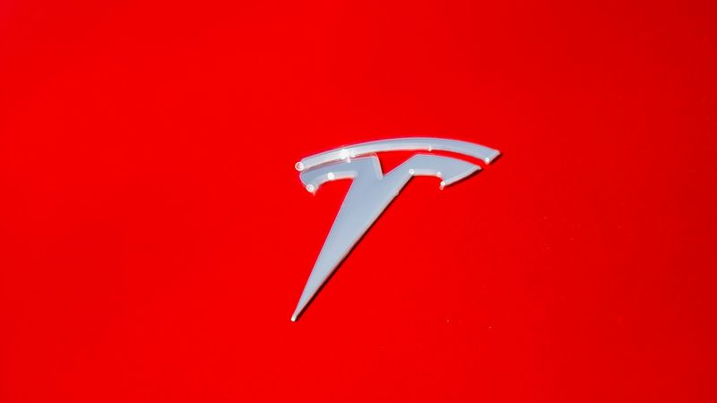 The image features the Tesla logo prominently displayed against a bright red background. The logo, known for its sleek and modern design, symbolizes the electric vehicle company's innovative approach to automotive technology.