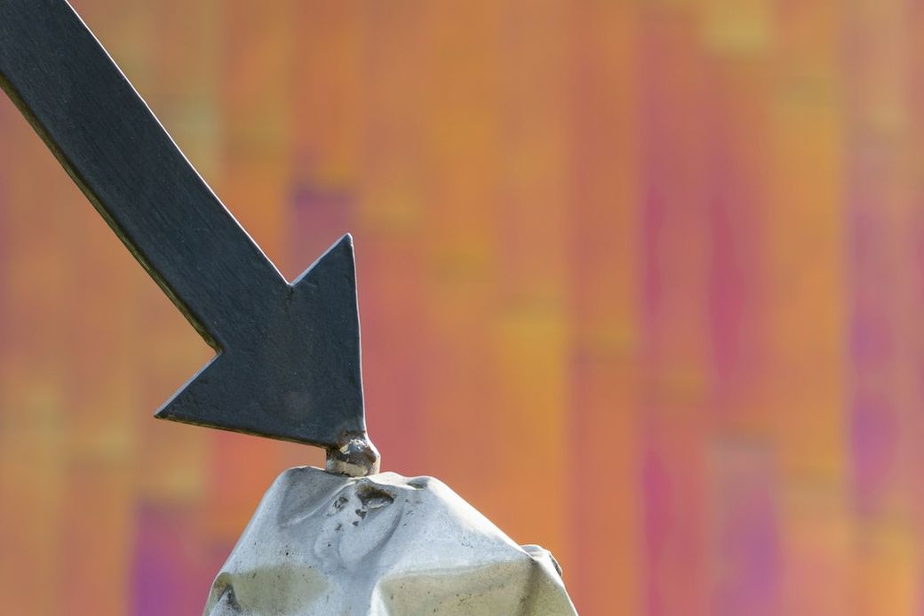 The image features a large black arrow pointing downwards, tipped on a gray rock formation. The background shows an abstract blurred pattern with warm colors, creating a stark contrast with the arrow and rock. — Credit: Photo by Fer Troulik on Unsplash