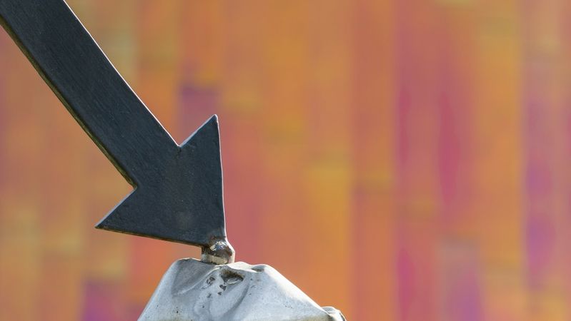 The image features a large black arrow pointing downwards, tipped on a gray rock formation. The background shows an abstract blurred pattern with warm colors, creating a stark contrast with the arrow and rock.