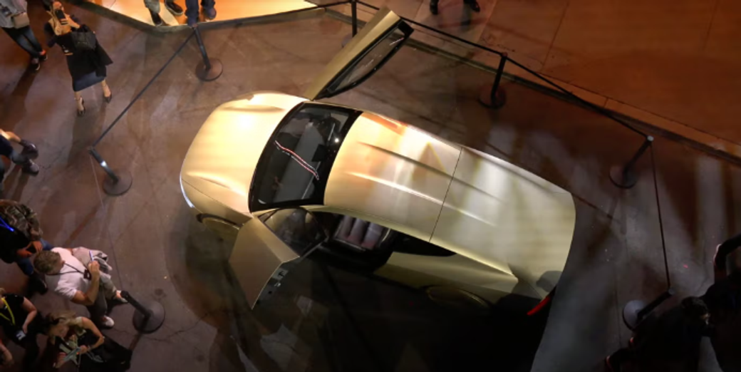 The image shows a sleek, futuristic vehicle with its doors open, displayed in an outdoor environment surrounded by barriers. A crowd of people can be seen around it, engaging with the design, which suggests an event showcasing innovative automotive technology, possibly linked to Tesla's developments in autonomous vehicle designs.