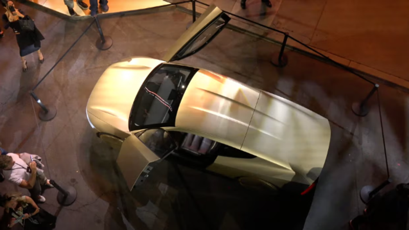 The image shows a sleek, futuristic vehicle with its doors open, displayed in an outdoor environment surrounded by barriers. A crowd of people can be seen around it, engaging with the design, which suggests an event showcasing innovative automotive technology, possibly linked to Tesla's developments in autonomous vehicle designs.