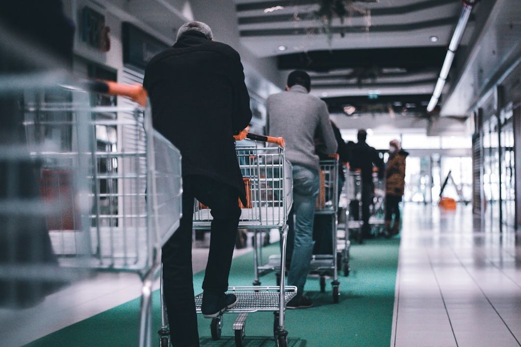 The image shows a line of shoppers pushing shopping carts down a corridor in a retail space. The view focuses on the backs of the shoppers as they navigate the smooth, tiled floor. — Credit: Photo by Adrien Delforge on Unsplash
