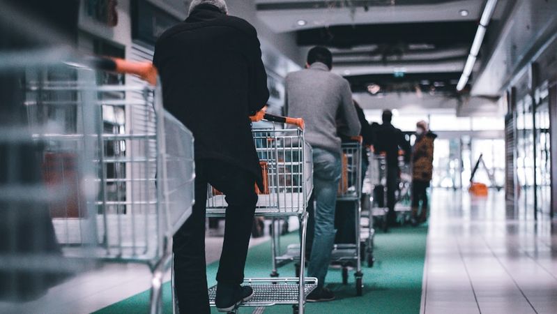The image shows a line of shoppers pushing shopping carts down a corridor in a retail space. The view focuses on the backs of the shoppers as they navigate the smooth, tiled floor.