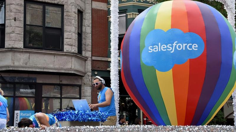 The image captures a vibrant parade scene featuring a DJ performing on a float, accompanied by a large colorful hot air balloon displaying the Salesforce logo. The atmosphere is festive and lively, indicative of a community celebration or event.