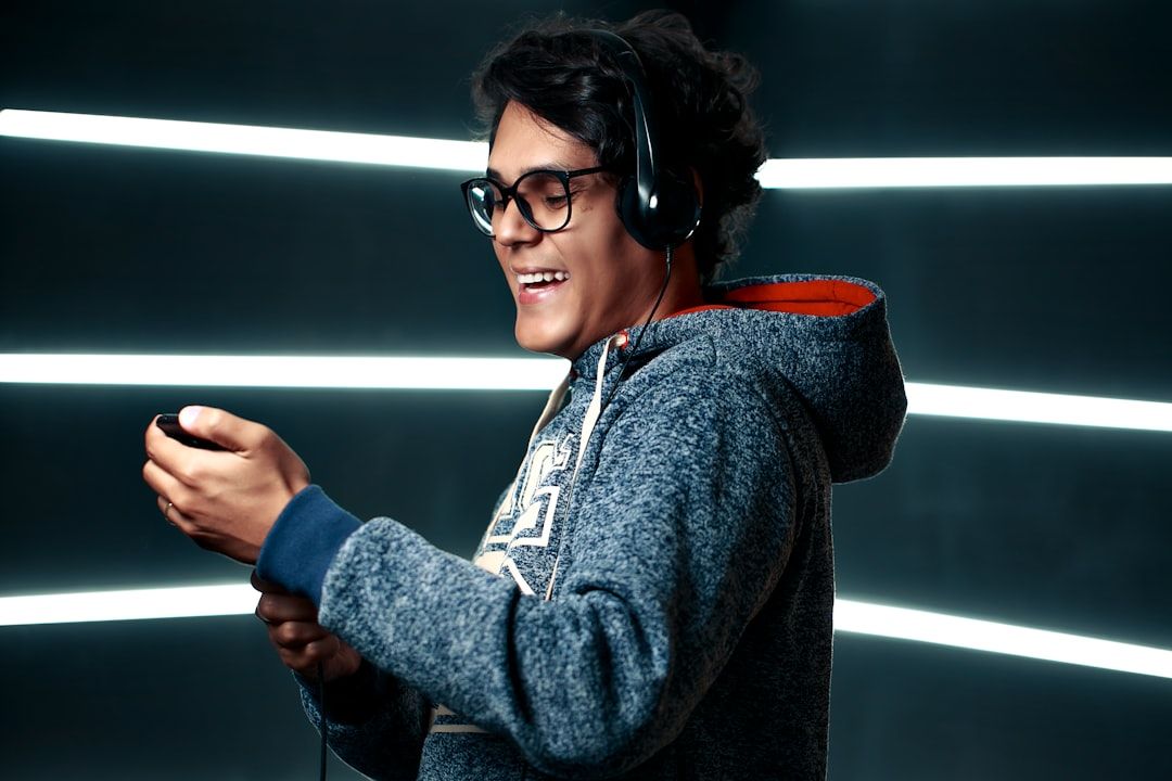 A young man wearing glasses and a hoodie is enjoying music with headphones on. He is smiling while holding a music device, set against a backdrop of illuminated lines.