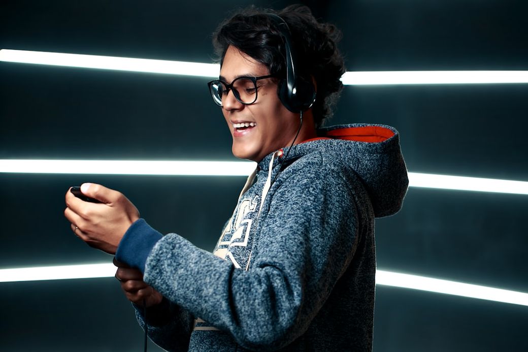 A young man wearing glasses and a hoodie is enjoying music with headphones on. He is smiling while holding a music device, set against a backdrop of illuminated lines. — Credit: Photo by Fábio Lucas on Unsplash