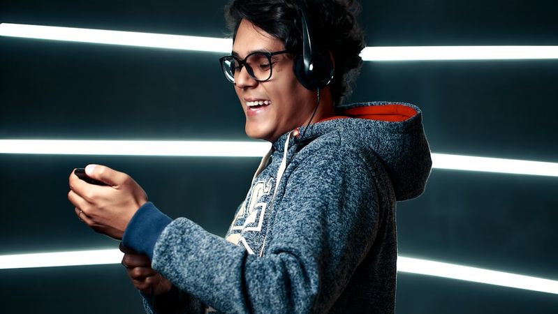A young man wearing glasses and a hoodie is enjoying music with headphones on. He is smiling while holding a music device, set against a backdrop of illuminated lines.