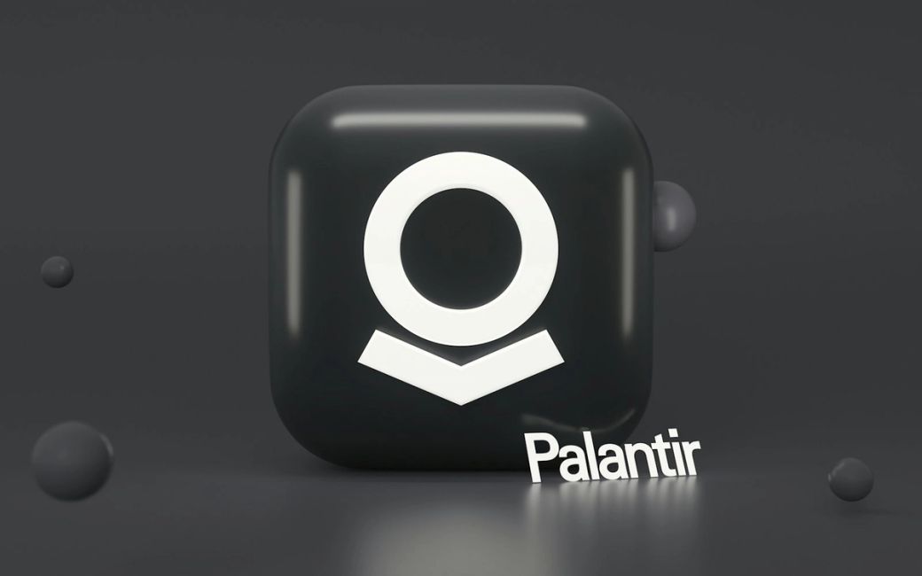 The image features a three-dimensional representation of the Palantir logo on a dark background. Accompanying the logo is the text 'Palantir' displayed prominently in a modern font. — Credit: Photo by Mariia Shalabaieva / Unsplash
