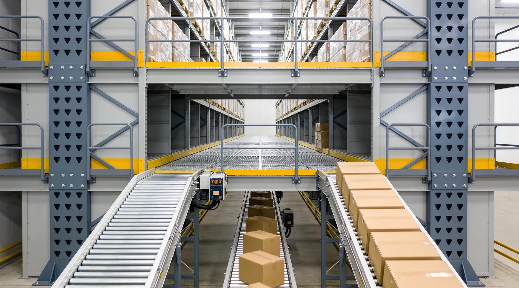 Multi-level rack-supported pick module with roller conveyor lines running through the ground level, elevated bar-grating walkways with grey guardrails and safety-yellow kick plates above, and pallet storage in the racking overhead