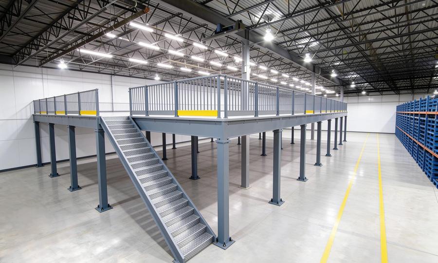 Free-standing steel mezzanine occupying part of an otherwise open warehouse floor, its regular grid of slate-grey square tube columns on bolted base plates standing inside yellow-striped floor markings, with an open deck edged by picket guardrails and yellow kick plates and a straight galvanized stair projecting out onto the slab