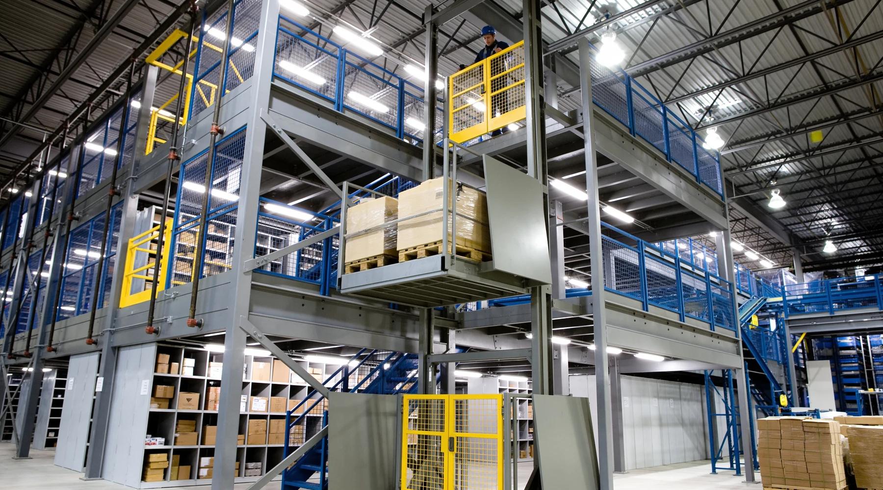 Caged vertical reciprocating conveyor lifting a wrapped pallet between floor and mezzanine deck with safety gates at both landings