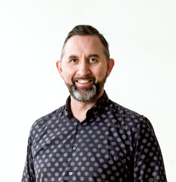 A light-skinned Māori man with energetic eyes framed by heavy eyebrows, wearing a salt and pepper beard, with short cropped hair and a blue/black polkadot shirt. He is smiling.