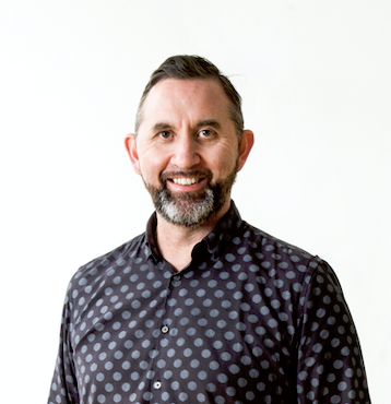 A light-skinned Māori man with energetic eyes framed by heavy eyebrows, wearing a salt and pepper beard, with short cropped hair and a  blue/black polkadot shirt. He is smiling.