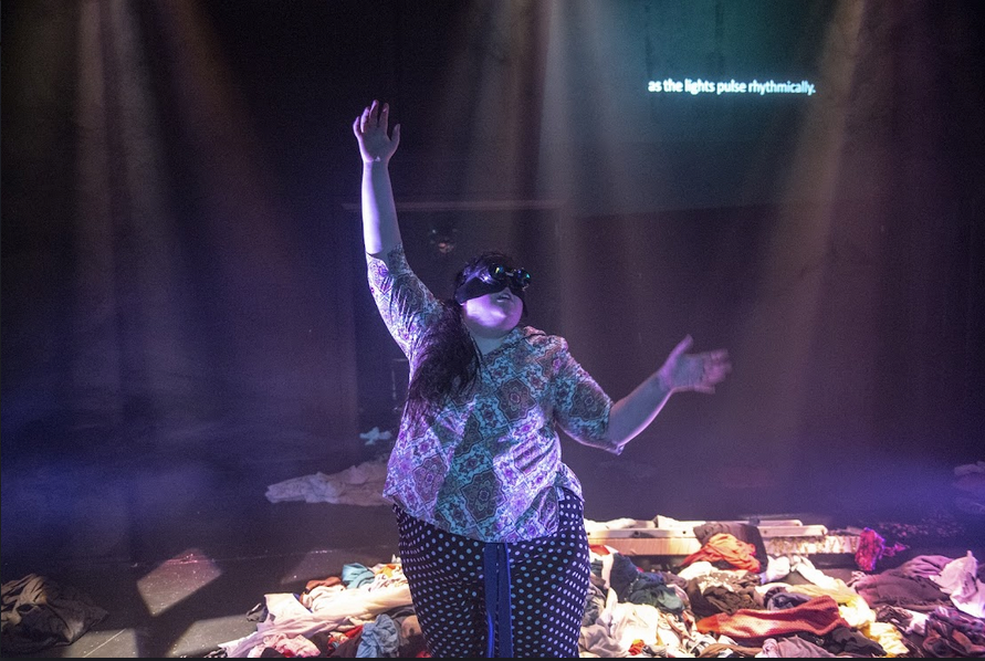 Susan Williams in the centre of the frame, with long dark hair swept to one side, a black bandana under aviator goggles, in the midst of a pile of laundry. Behind them is a screen with the projections "as the lights pulse rhythmically"