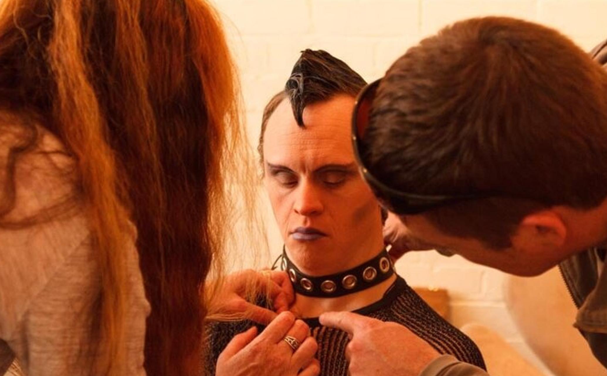 A Pākehā (Duncan) is sitting with two people in front of him fixing the front of his. top. He is wearing a collar, with purple lipstick on and his hair gelled forward into a spike.